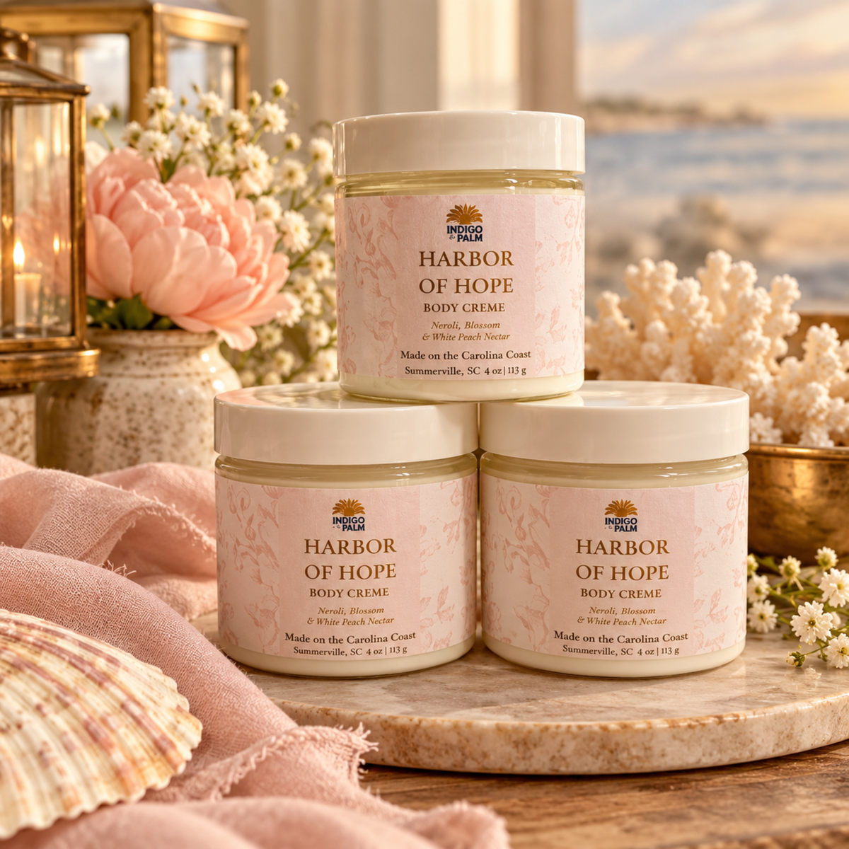 Three jars of Harbor of Hope Body Creme stacked on a marble board with pink fabric, flowers, and a beach backdrop.