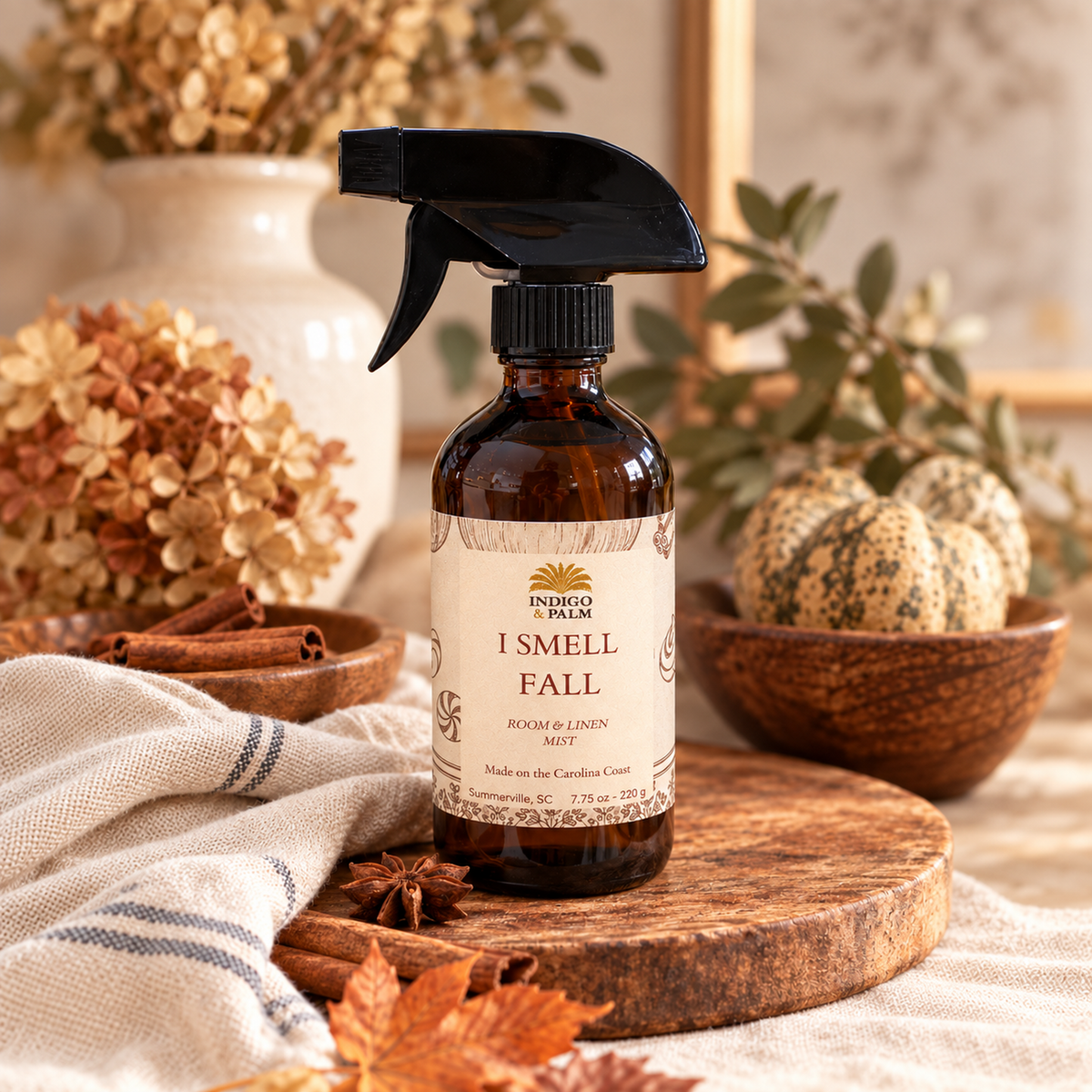 An amber glass spray bottle with a black trigger sprayer and a cream label reading 'I SMELL FALL' by Indigo & Palm, styled on a wooden board with dried flowers, a striped cloth, and autumn decor in a warm indoor setting.